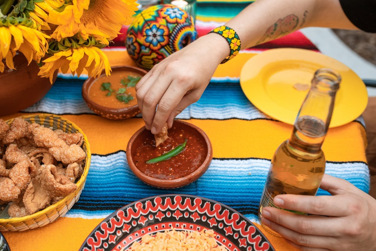 A colorful Mexican spread featuring salsa, chicharrones, and beer on a striped tablecloth.