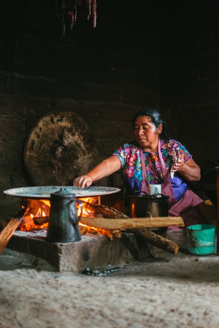 Indigenous woman cooking over an open flame in a rustic Mexican setting.